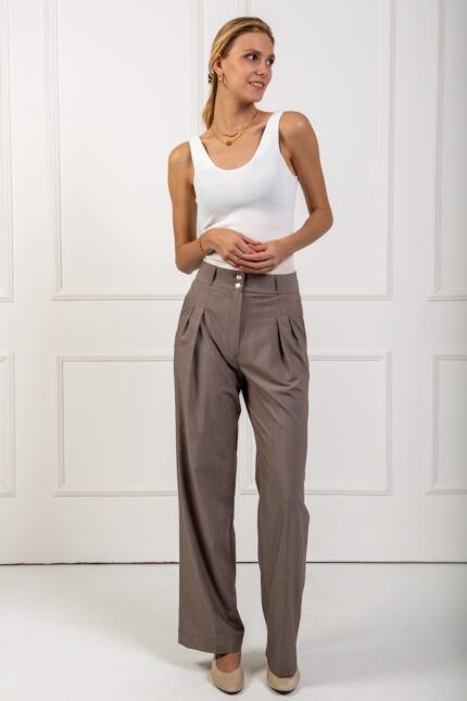 The blonde girl wears wide brown trousers and a white top, standing in front of a white wall.
