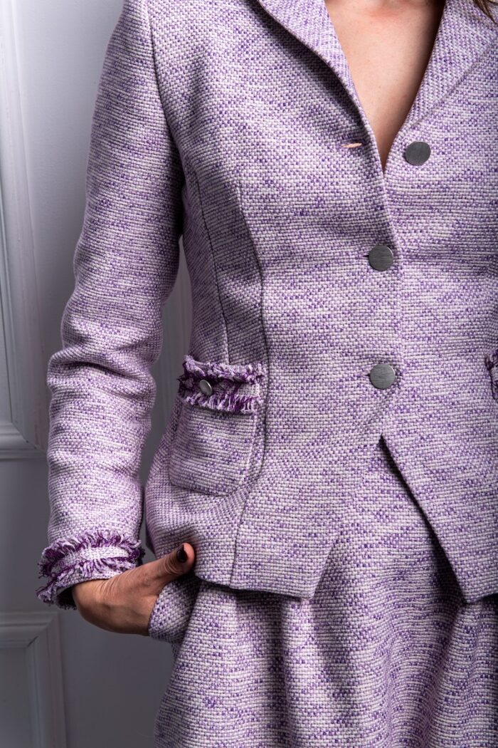 The woman wears a purple cotton blazer with small lapels. He has his hands in his pockets and is standing in front of a white wall.