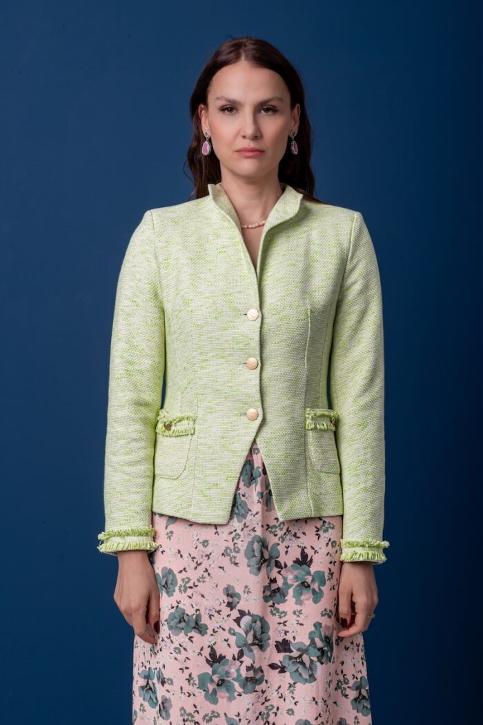 Žena nosi zeleni pamučni blejzer i floralnu haljinu i stoji ispred plave pozadine.